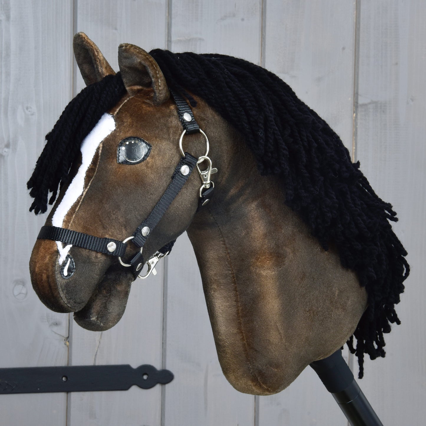 The Hobby Horse LarDen "Hobby Horse Sundance with Black Halter" features a brown head inspired by the swiftness of stallions, with a black yarn mane, white blaze, and black halter, all mounted on a sturdy wooden stick in front of a gray wooden wall.