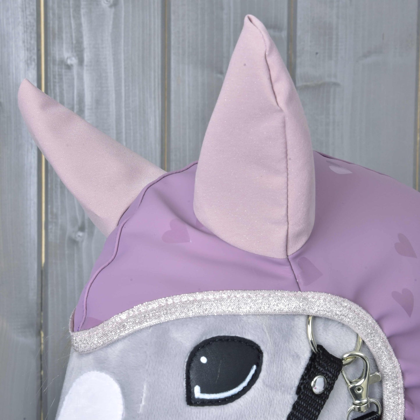 A close-up of the Hobby Horse LarDen "Raincoat Sweetheart" plush horse head with light purple ears and a heart-patterned cover, set against a wooden background—a charming all-weather hobby horse accessory.