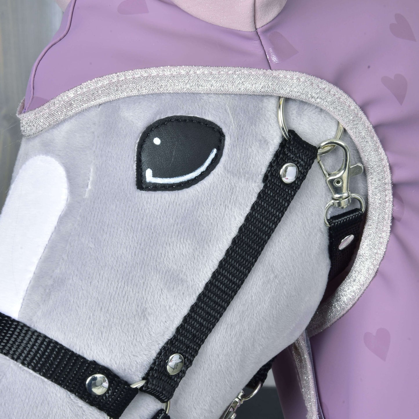 A close-up of Hobby Horse LarDen’s plush gray hobby horse in a black bridle, wearing the Raincoat Sweetheart accessory, beside a person in a lavender heart-patterned outfit. The horse has a stitched smiling eye and glittery neck trim.