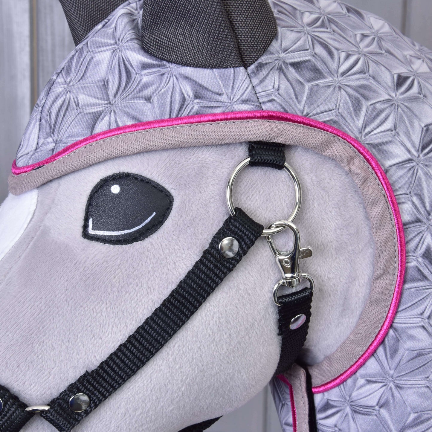 Close-up of Hobby Horse LarDen's Raincoat Valentine, a plush horse’s head wearing a geometric-patterned gray fabric with black bridle and pink trim, featuring a metal ring clip—an all-weather accessory for stylish protection.