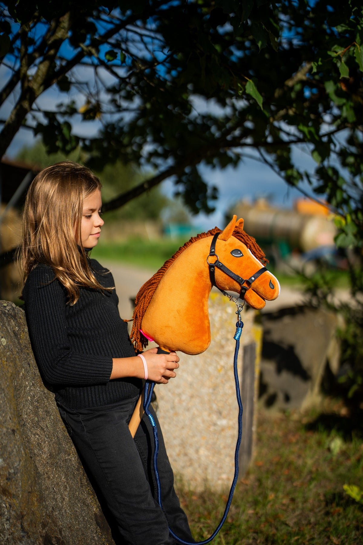 Dziewczyna w czarnej bluzce opiera się o kamienny blok pod drzewem, trzymając Hobby Horse Wendy z kantarem Black Halter od Hobby Horse LarDen – jasna kasztanowata pluszowa głowa, ruda grzywa z włóczki – pokazany z Uwiązem Blues rope stripe