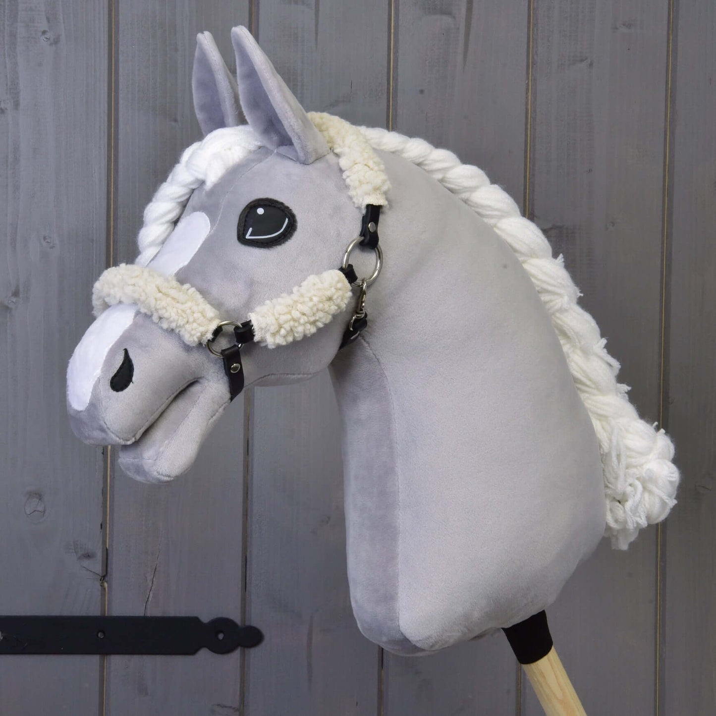 Hobby Horse LarDen przedstawia pluszowego szarego Hobby Horse z czarnym kantarem z pełnym futerkiem i elementami ze skóry ekologicznej, osadzonego na drewnianym kiju na tle drewnianych desek.